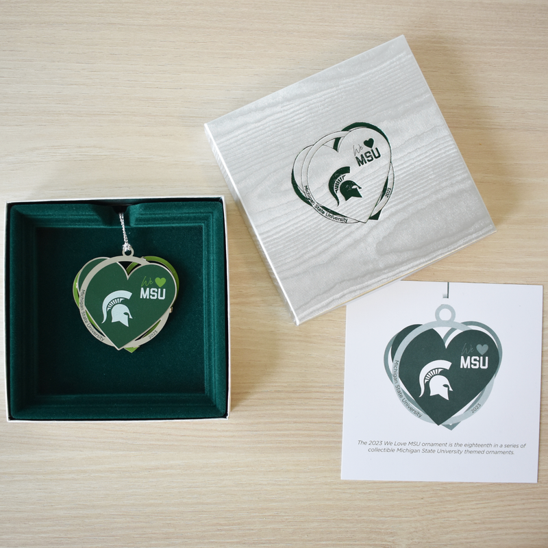 A silver ornament made of three separate layers features a bold white Spartan helmet with text reading "we 'heart' MSU" on the upper right quadrant of a green heart. The silver ring layer reads Michigan State University 2023. The ornament is nestled in its silver box with a felt green lining, and is placed next to the silver box top foil embossed in forest green and the matching insert card.
