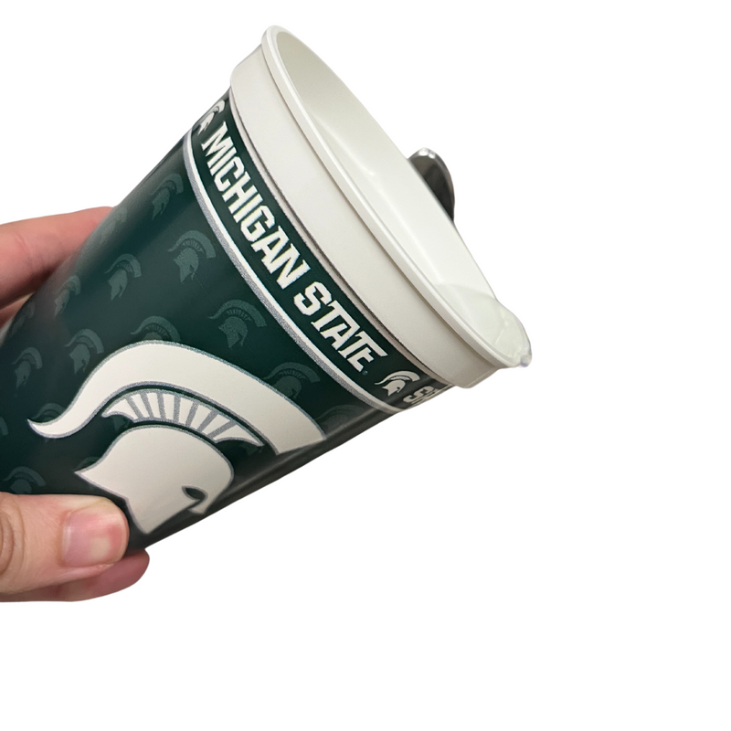 A person pouring water out of a plastic 16 oz. cup, featuring a wraparound design that includes a large white Spartan helmet in the center with a dark green background with faded green Spartan helmet pattern. Around the rim it reads Michigan State Spartans.