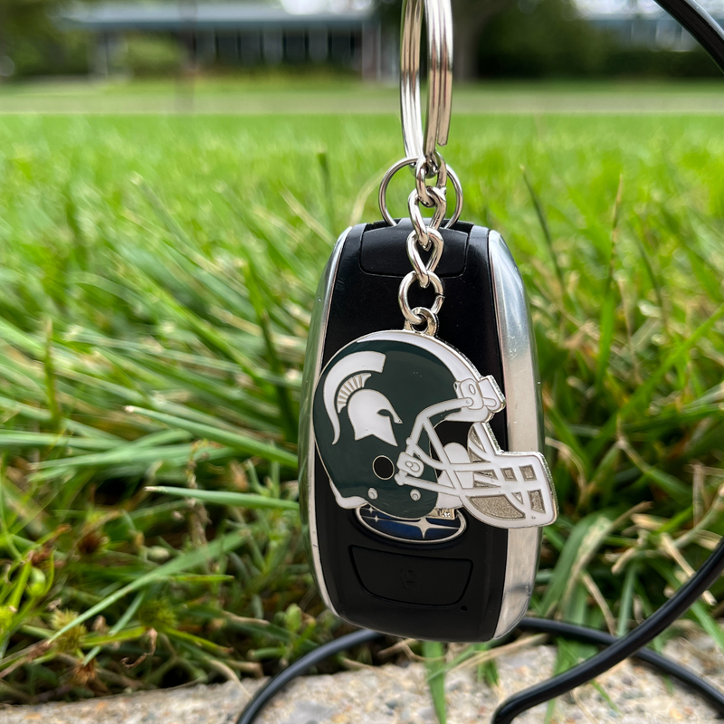 Silver metal is cutout in the shape of a football helmet and enameled in dark green and white following the MSU classic helmet style. Attached by a silver chain is a detachable keyring.