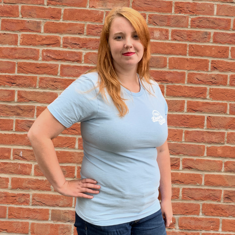 A woman standing in front of a brick wall wearing a light blue t-shirt  with an MSU Dairy Store logo on center chest.