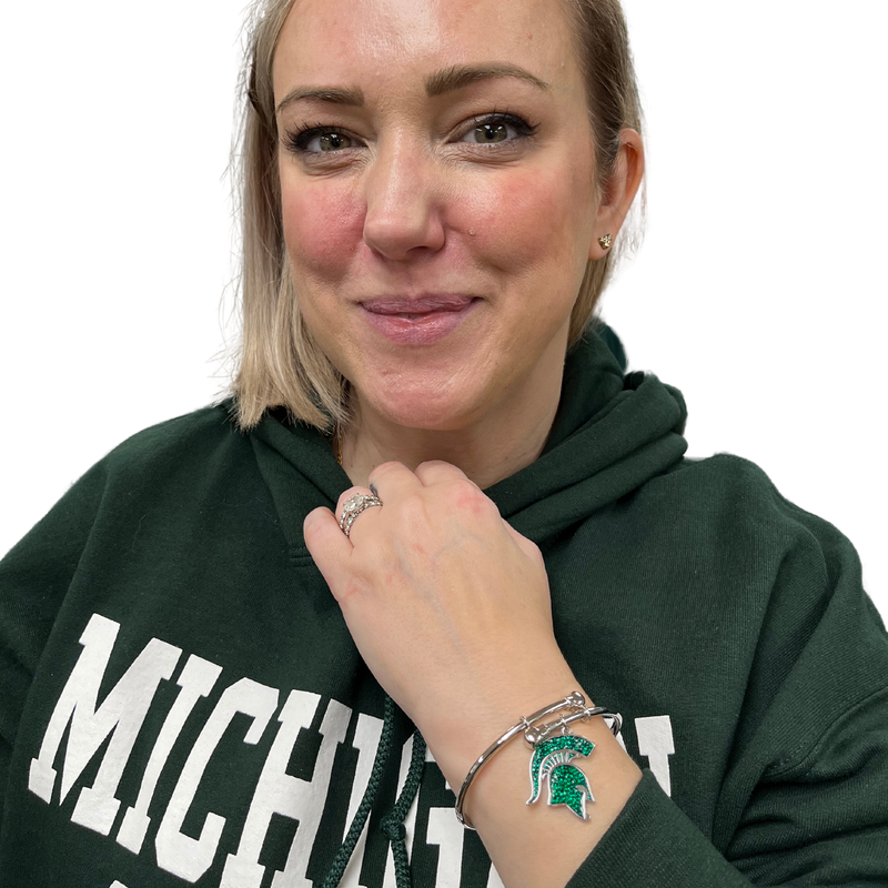 A woman modeling a silver, bangle style bracelet with a Michigan State spartan helmet logo medallion that is filled with green crystals.