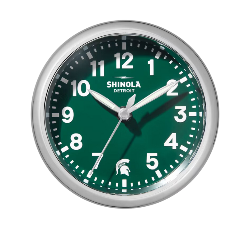 Chrome clock with green face and white lettering. Shinola Detroit displayed under the 12 and a Spartan helment in the place of the 6. White lettering for the hours and minutes, with chrome sweeping hands.