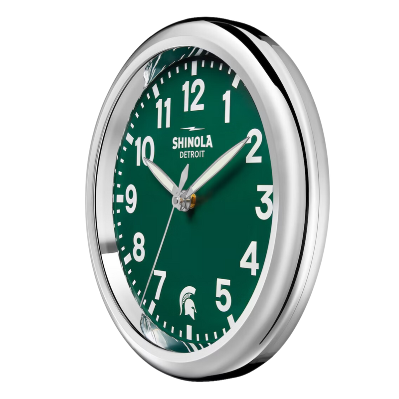 Chrome clock with green face and white lettering. Shinola Detroit displayed under the 12 and a Spartan helment in the place of the 6. White lettering for the hours and minutes, with chrome sweeping hands.