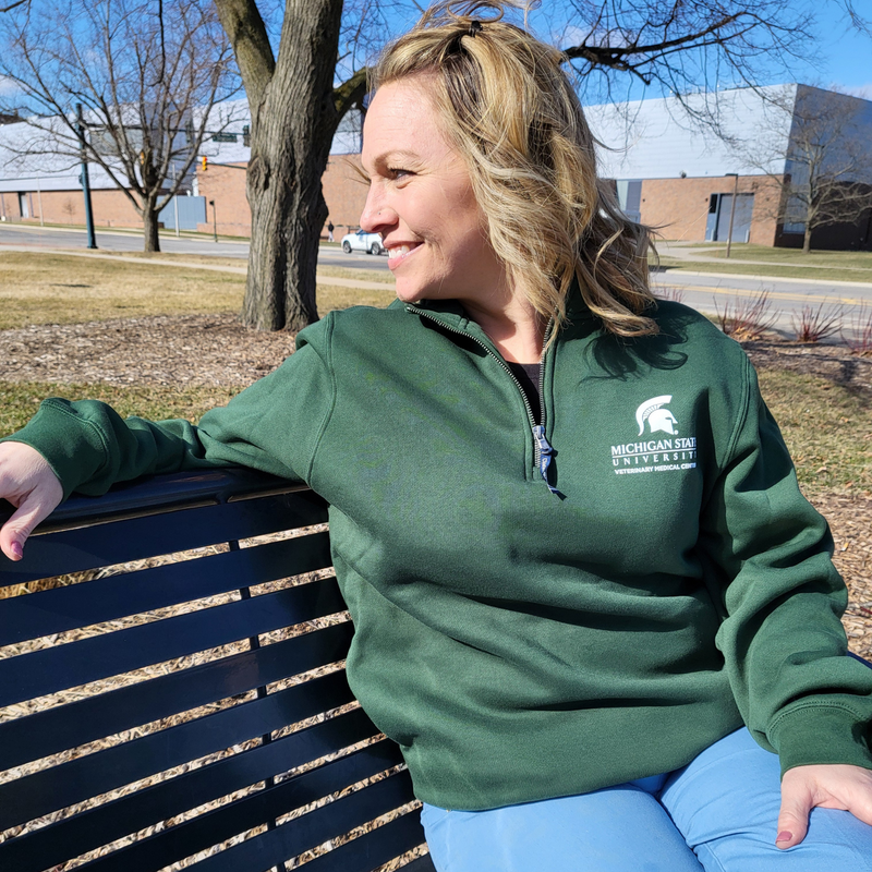 A woman in blue scrubs and a dark green, quarter zip sweatshirt sitting on a park bench looking over her right shoulder. The upper left chest is embroidered with a Veterinary Medical Center logo in white.