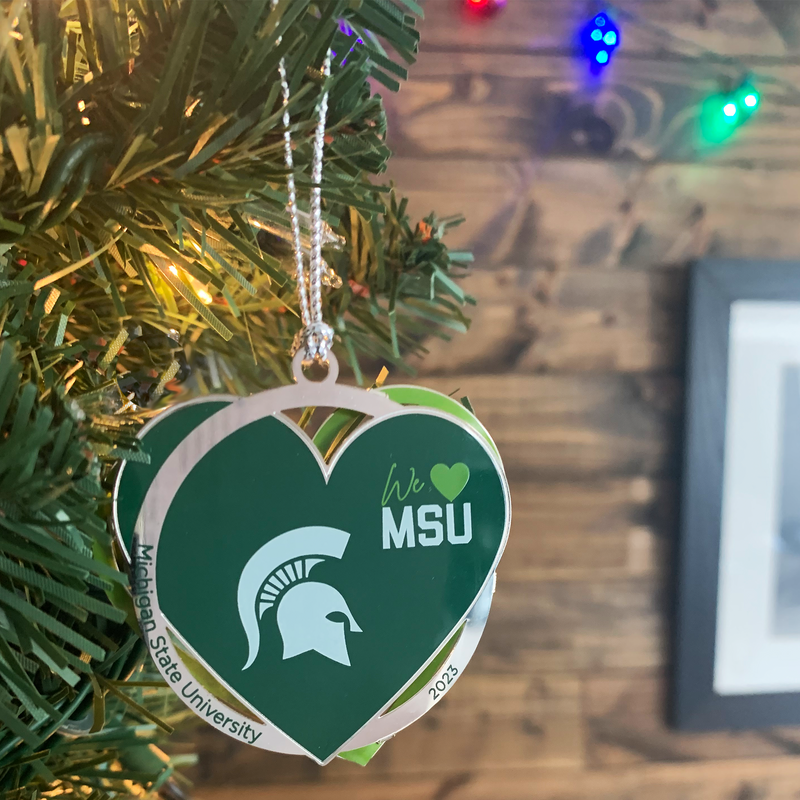 A silver ornament made of three separate layers features a bold white Spartan helmet with text reading "we 'heart' MSU" on the upper right quadrant of a green heart. The silver ring layer reads Michigan State University 2023. The ornament is hanging from an artificial tree with colorful holiday lights on a shiplap wall in the background.