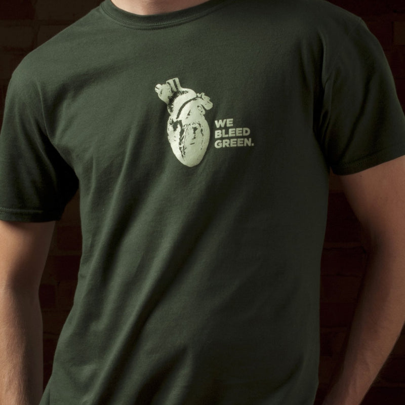 A person wearing a forest green short sleeve crewneck t-shirt with a digram of a heart and text reading We Bleed Green printed in white on the center chest.