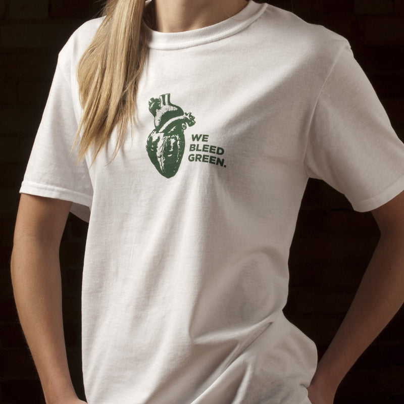 A person wearing a white short sleeve crewneck t-shirt with a digram of a heart and text reading We Bleed Green printed in forest green on the center chest.