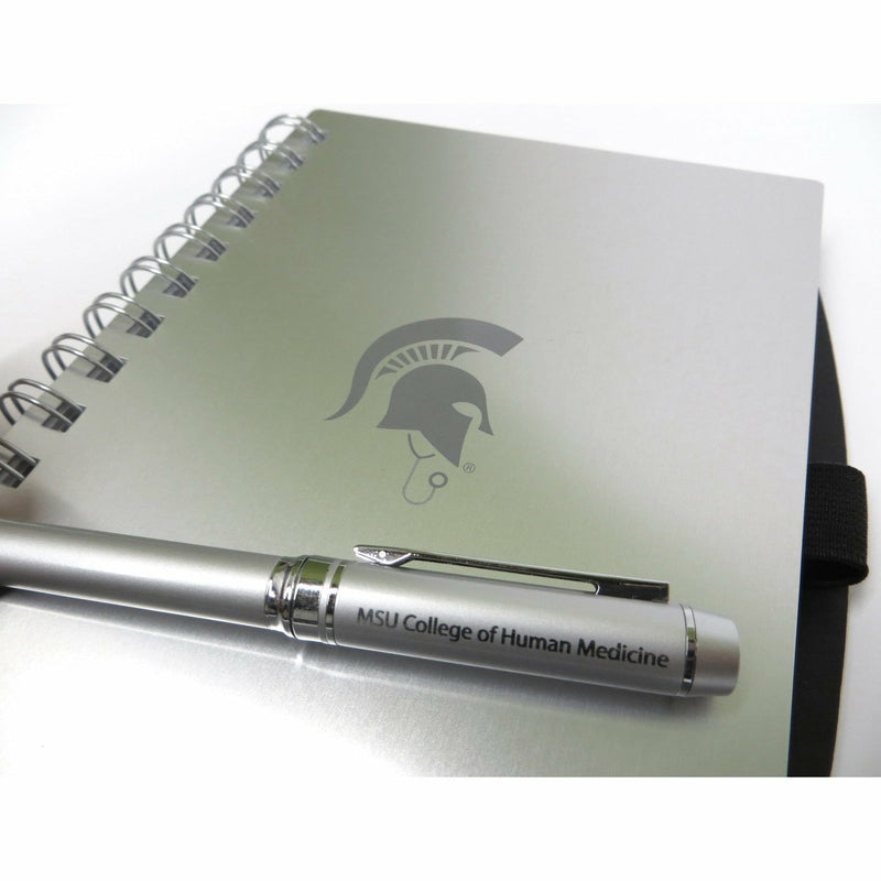 Close-up of the top of the silver pen which reads MSU College of Human Medicine, laying atop the Spartan helmet insignia on the notebook