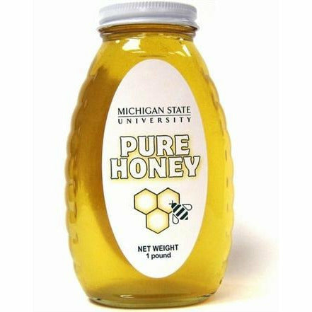 Clear glass jar with a slight texture for grip on the sides, filled with golden honey. White label in the middle includes the MSU wordmark, text reading Pure honey: net weight 1 pound, and an illustrated bee and honeycomb.
