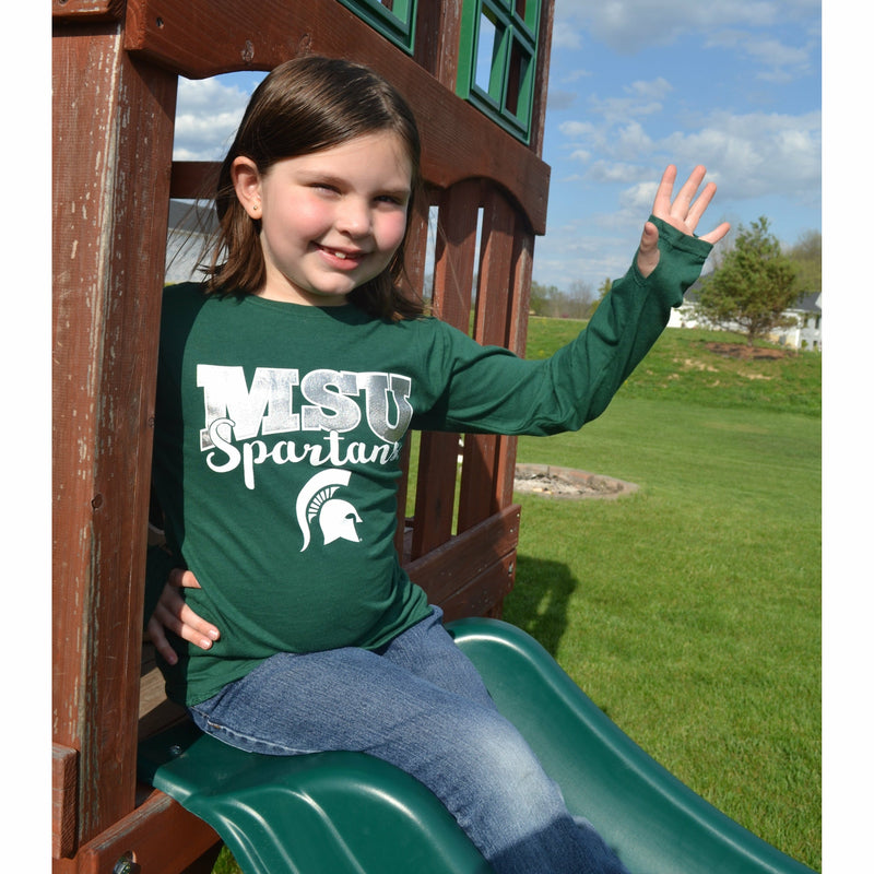 A young girl sits atop a slide playset waving her left hand to show the thumb hole design of the Youth Long Sleeve t-shirt