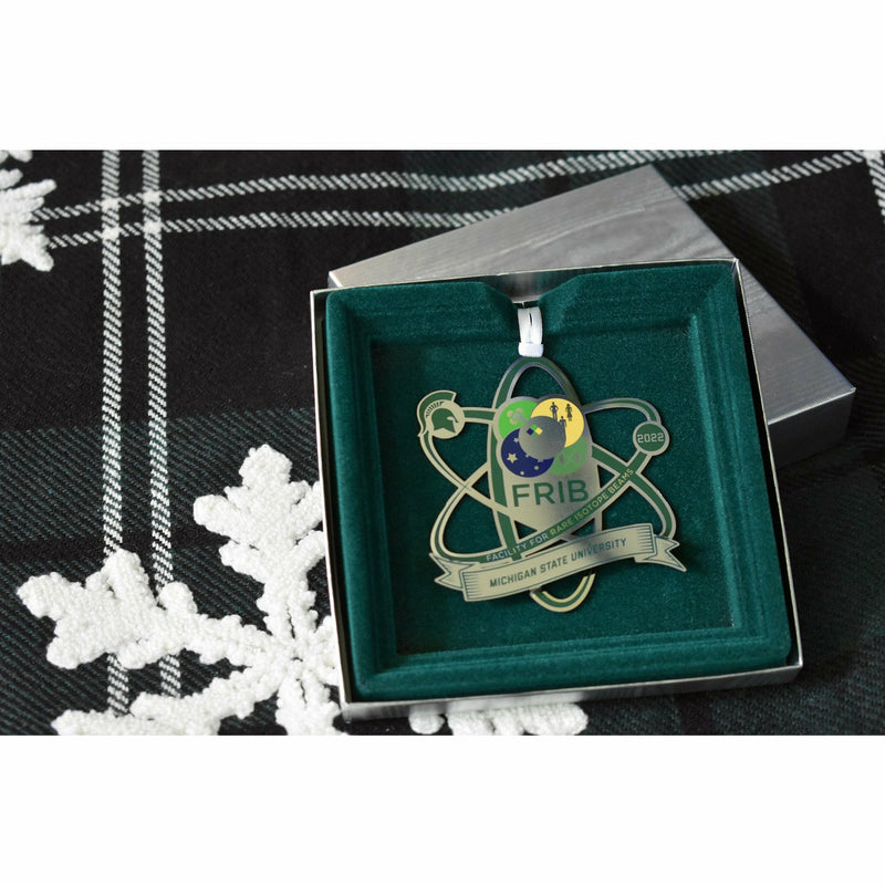 Top-down view of the FRIB 2022 Michigan State University ornament nestled in a silver box with a green velvet lining. Box is stacked on a plaid snowflake-patterned tree blanket. 
