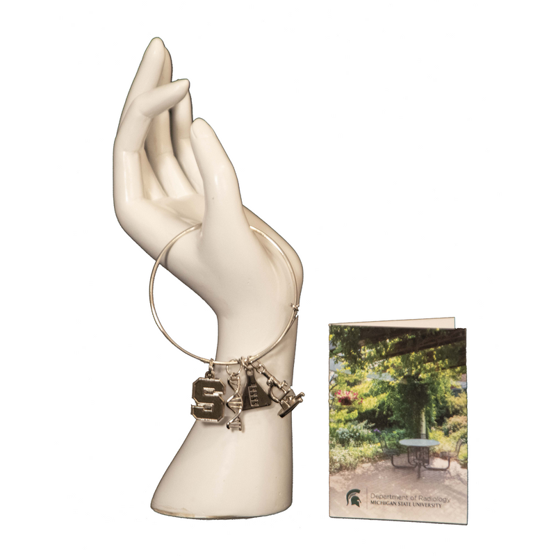 The silver supporting lab workers bracelet on a hand mannequin next to a greeting card featuring a photo of a picnic table under a pergola in the gardens.
