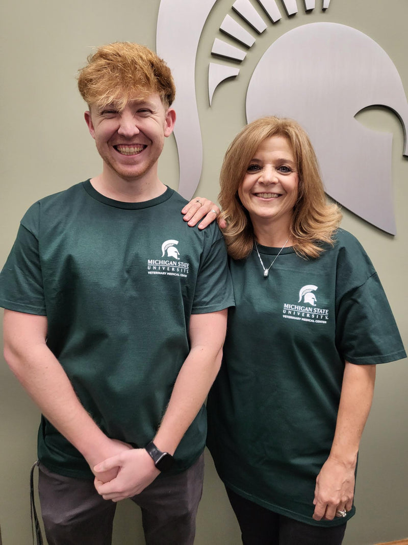 Two people with green t-shirts. Left upper side featuring the Veterinary Medical Center logo and Spartan helmet in white.