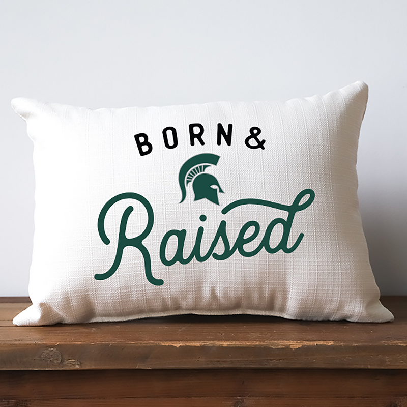 An off-white lumbar pillow with a thatched texture. Spanning across the front are the words "Born and Raised," with "Raised" in green cursive. Between the words is a Spartan helmet logo.