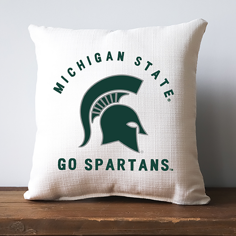 An off-white square pillow with a thatched texture. Centered on the front is a large green Spartan helmet outlined in gray. Arched above are block letters reading Michigan State. Below the helmet, larger block letters read “Go Spartans"