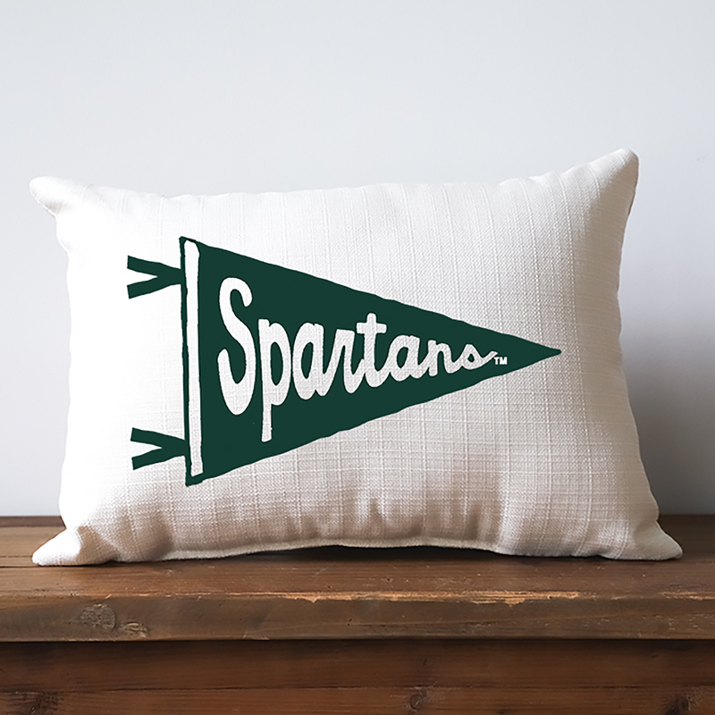 An off-white lumbar pillow with a thatched texture. Spanning across the front is an illustrated green pennant flag with cut-out script font reading Spartans.