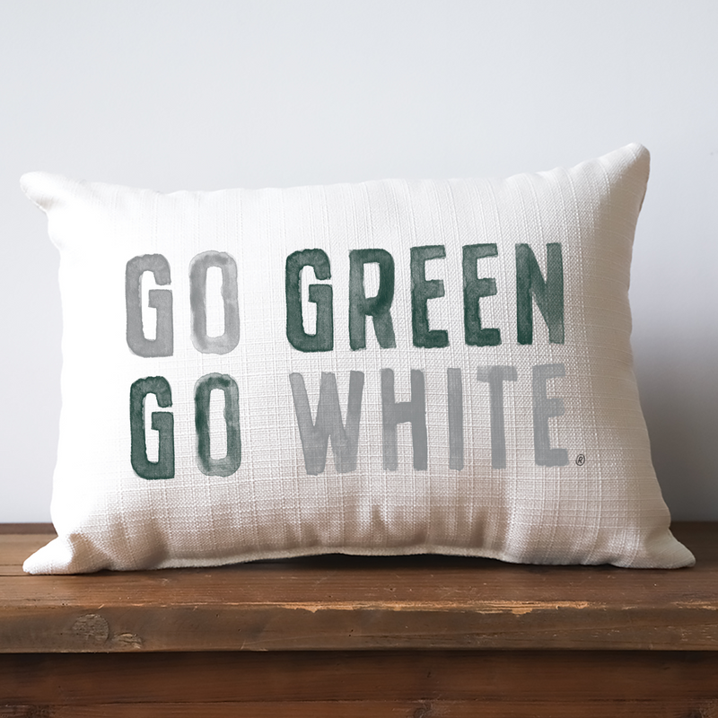 An off-white lumbar pillow with a thatched texture. Spanning across the front are watercolor block letters spelling Go Green, Go White, with each word alternating between light gray and forest green