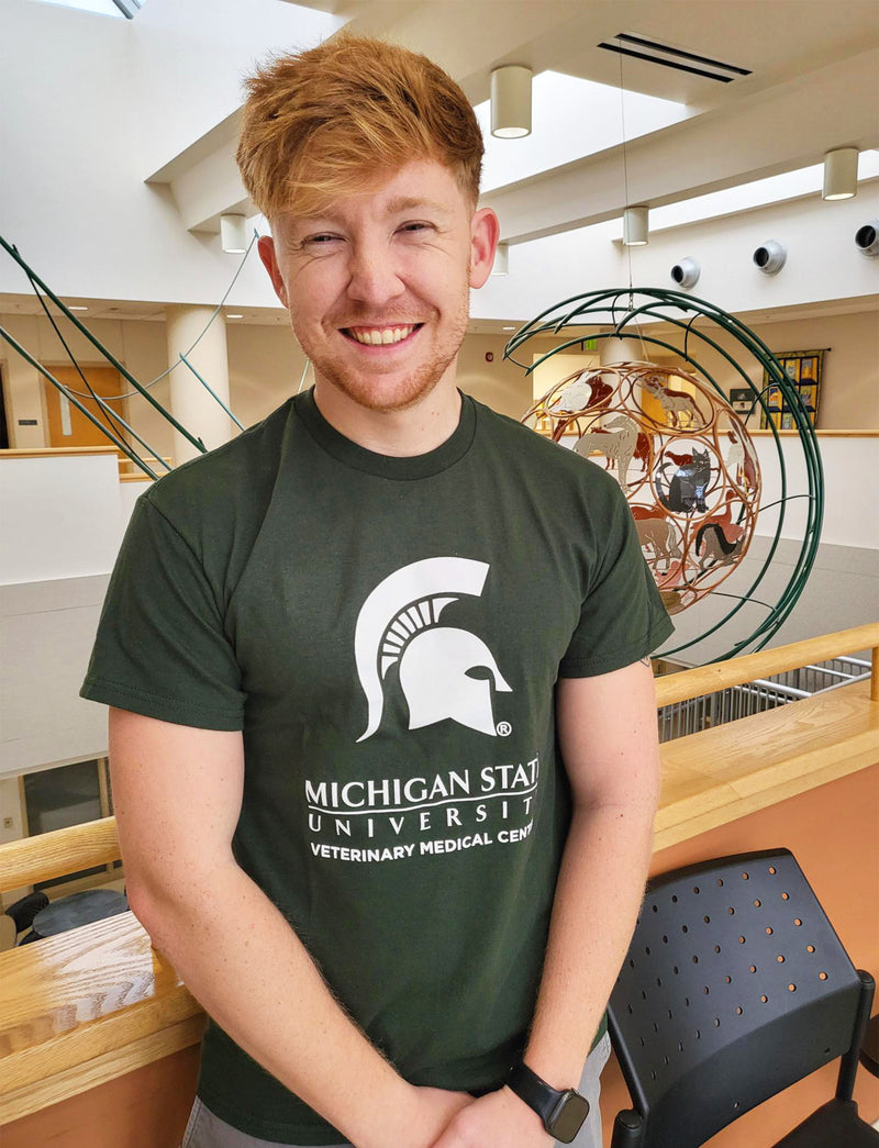 A man in blue jeans wearing a green short-sleeve t-shirt. Across the center chest is a large white Spartan helmet centered above the Michigan State wordmark logo. Beneath that is large block text reading “Veterinary Medical Center.”