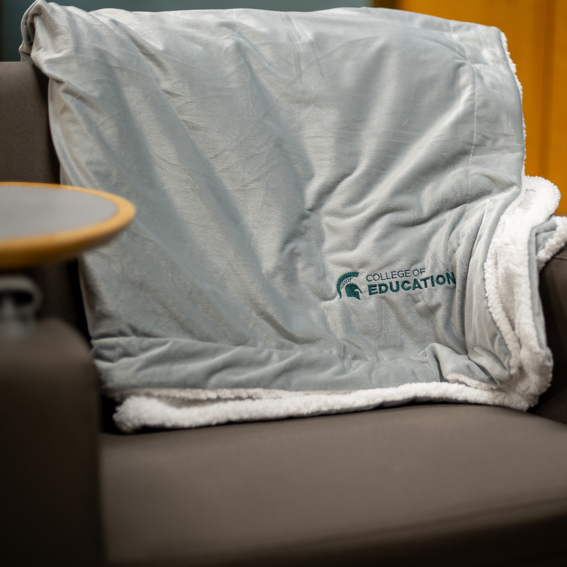 A gray plush blanket with a white, sherpa fleece lining. On the bottom right corner of the blanket, engraved in green, is a MSU spartan helmet logo with the words "College of Education". The blanket is sitting on a brown office chair. 