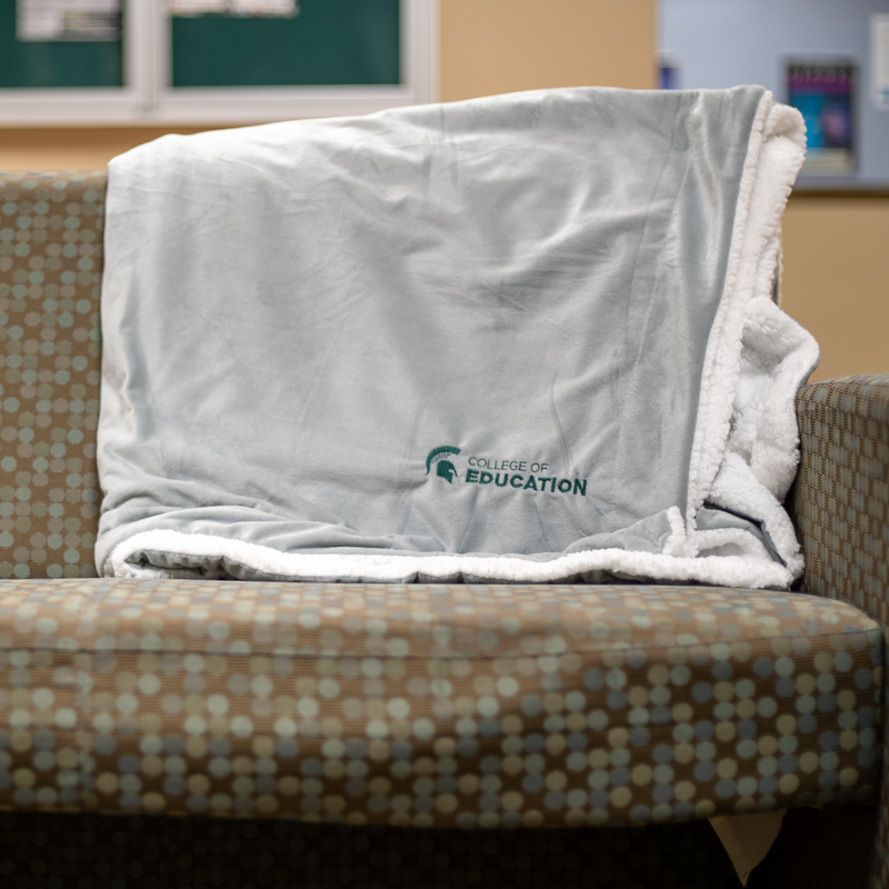 A gray plush blanket with a white, sherpa fleece lining. On the bottom right corner of the blanket, engraved in green, is a MSU spartan helmet logo with the words "College of Education". The blanket is sitting on a multi-colored cushioned chair. 