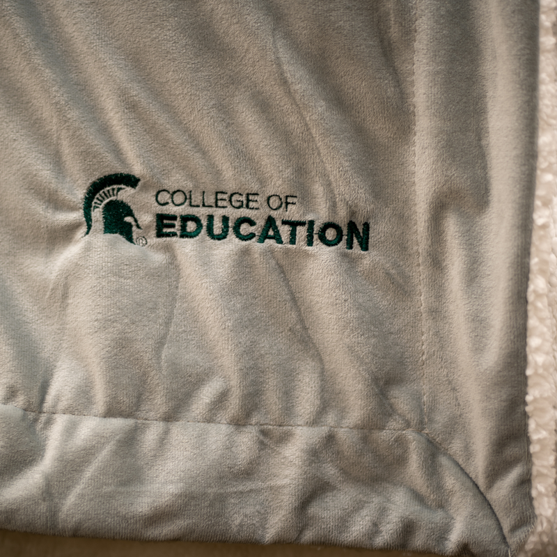 A gray plush blanket with a white, sherpa fleece lining. On the bottom right corner of the blanket, engraved in green, is a MSU spartan helmet logo with the words "College of Education".
