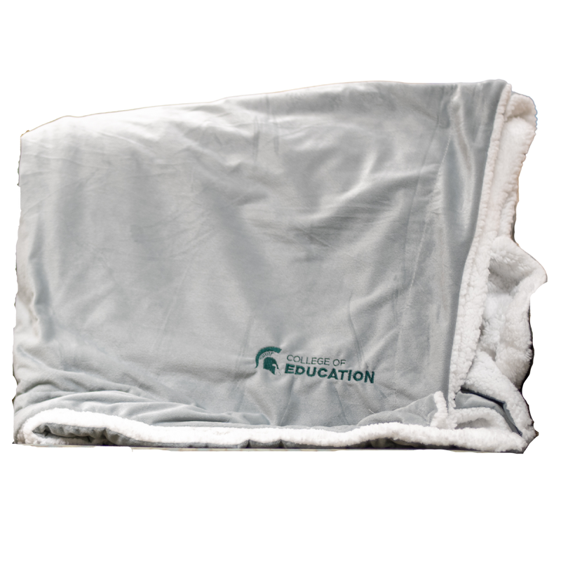 A gray plush blanket with a white, sherpa fleece lining. On the bottom right corner of the blanket, engraved in green, is a MSU spartan helmet logo with the words "College of Education". 