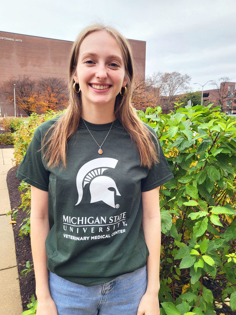 A woman in blue jeans wearing a green short-sleeve t-shirt. Across the center chest is a large white Spartan helmet centered above the Michigan State wordmark logo. Beneath that is large block text reading “Veterinary Medical Center.”