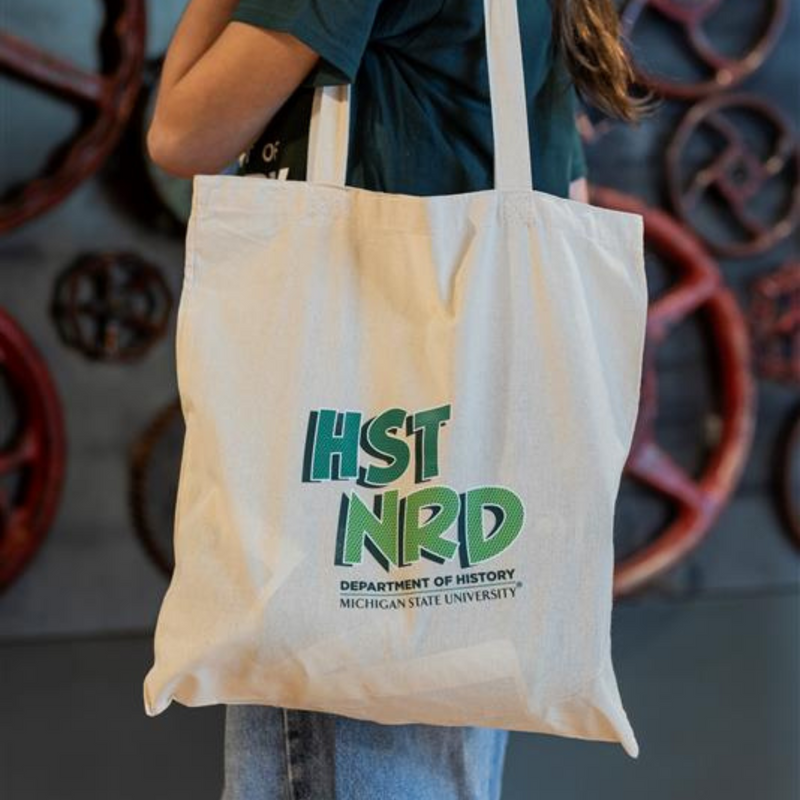 Person holding 7" x 9" natural cotton tote bag with ‘HST NRD’ above ‘Department of History’ and ‘Michigan State University’.