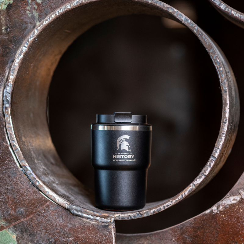 A black 12oz mug with a laser-etched Department of History logo and Spartan helmet photographed on a metal sculture.