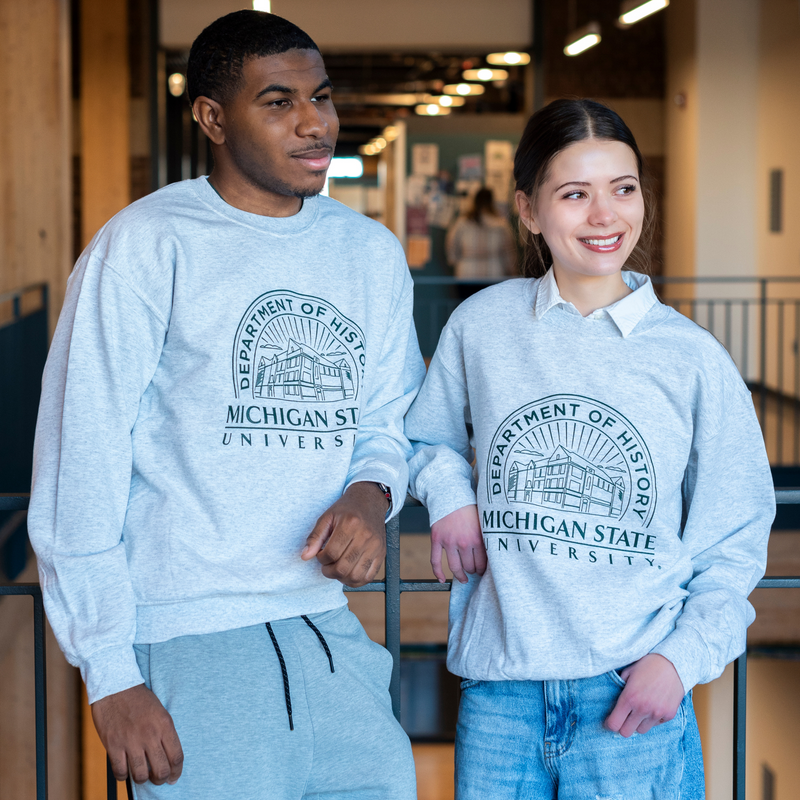 Two individuals wearing grey crewneck sweatshirts that showcases the Old Horticulture building surrounded by the text ‘Department of History’ and ‘Michigan State University’. 