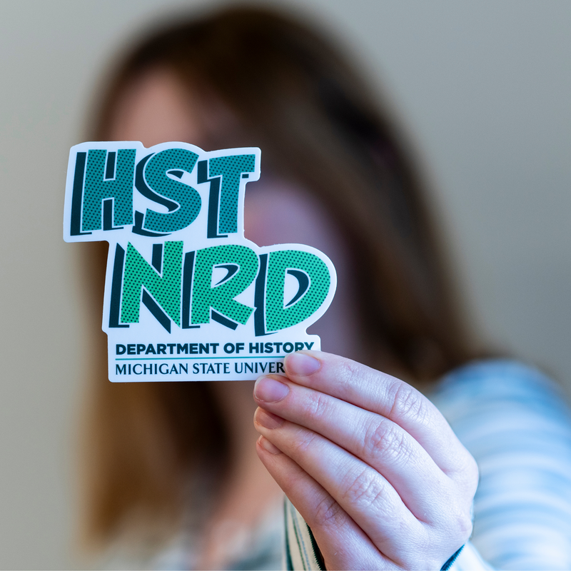 Person holding green and white vinyl sticker that features the text ‘HST NRD’ above of ‘Department of History’ and ‘Michigan State University’. 