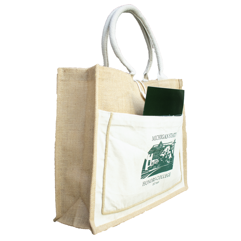 A green folder sticks out of the side of a tan and white tote bag made of jute and canvas material. On the side of the tote in green is a graphic of the Michigan State honors college. Above and below the image is the text "Michigan State Honors College Est. 1956"