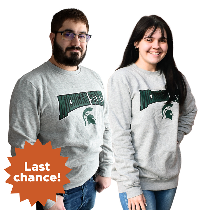 Man and woman standing side by side wearing a light heather gray crewneck sweatshirt, with Michigan State printed in a graphic arch over a Spartan helmet on the center chest. In the lower left corner is an orange sunburst with the words "Last chance!"