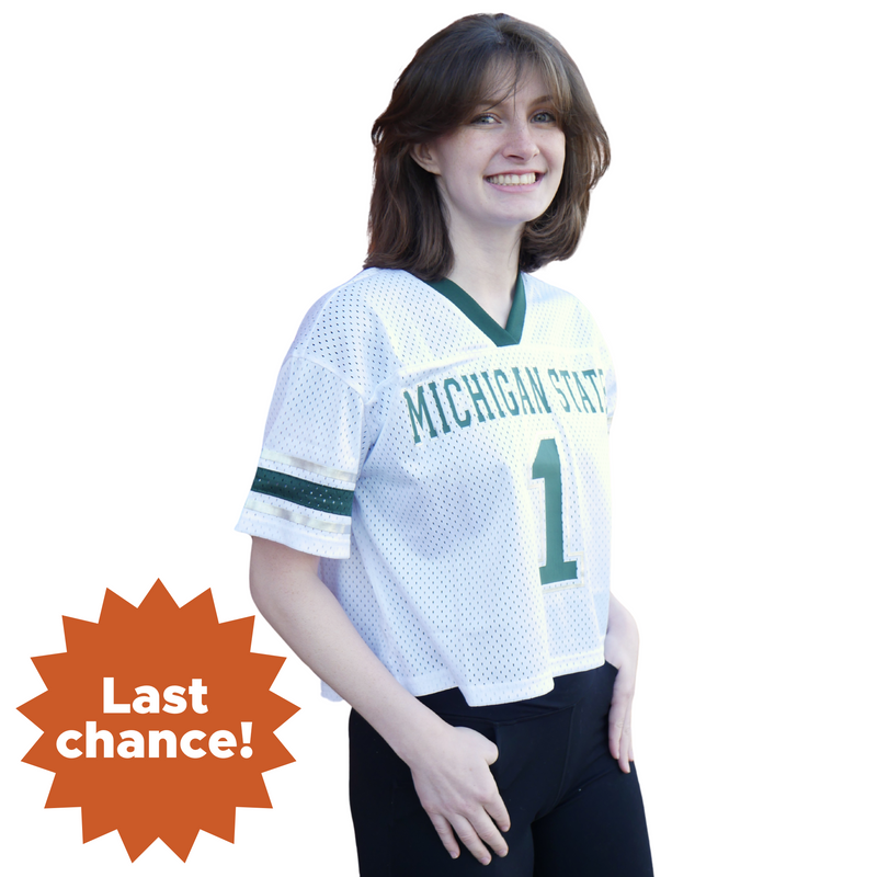 A woman wearing a white, cropped Michigan State jersey with a green neckline and sleeve cuffs. The torso has Michigan State written in all caps with the number one in green underneath. In the lower left corner is an orange sunburst with the words "Last chance!"