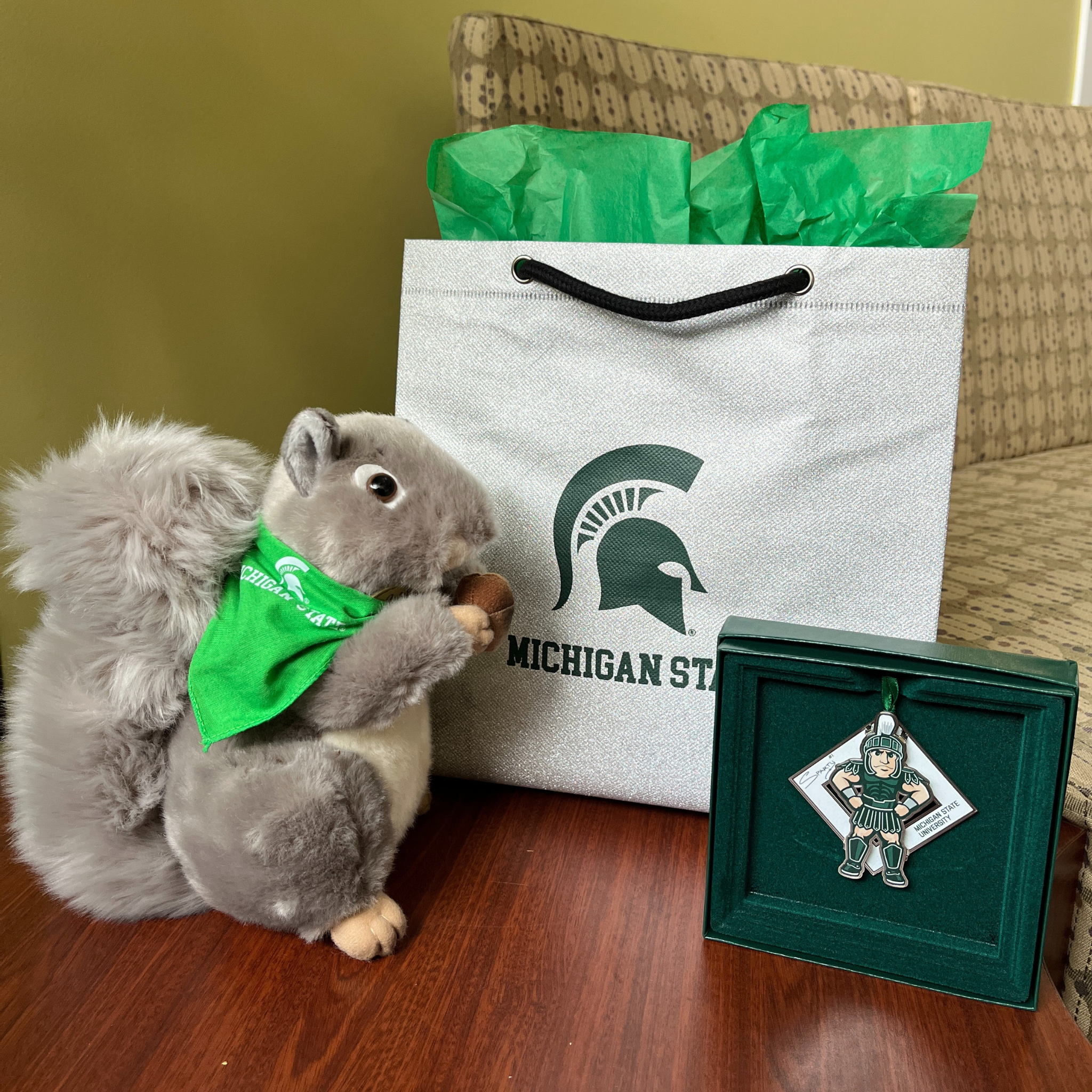 A plush squirrel wearing a green bandana and holding an acorn. To the right of the squirrel is a silver Michigan State gift bag with green tissue paper, and a 2024 MSU Sparty collectible ornament. 