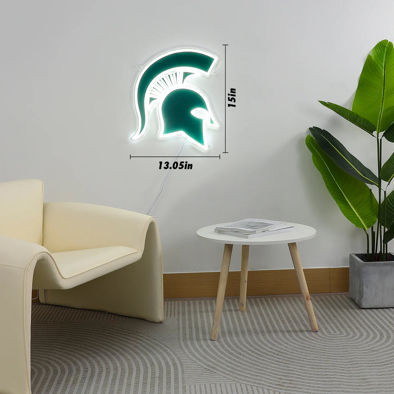 A neon wall sign that shows a green Michigan State Spartan helmet logo. Outlining the green logo are white neon lights. The sign measures 13.05 inches wide and 15 inches tall. It is hanging on a grey wall behind a tan chair a small three-legged desk. 