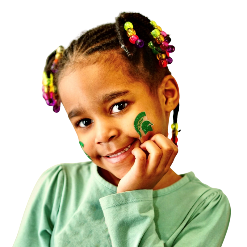 A little girl holds her hand up to a cheek, where a green glittery Spartan helmet body sticker is affixed.