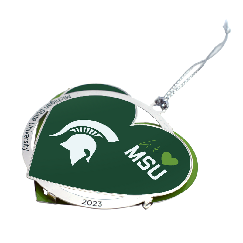 Close-up of the 2023 We Love MSU ornament at an angle. The bottom layer is a lime green outline of a heart. The top layer is a silver ring reading Michigan State University 2023 blended with a forest green solid heart. The heart features a white Spartan helmet to the left of center and in the upper right, text reads "We 'heart' MSU". A silver cord is tied to the top.