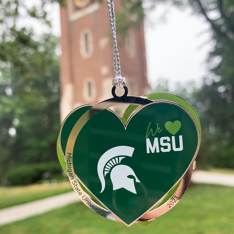 Close-up of the 2023 We Love MSU ornament at an angle. The bottom layer is a lime green outline of a heart. The top layer is a silver ring reading Michigan State University 2023 blended with a forest green solid heart. The heart features a white Spartan helmet to the left of center and in the upper right, text reads "We 'heart' MSU". A silver cord is tied to the top. Beaumont Tower is in the background.