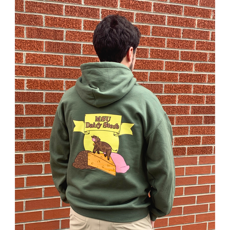 Man standing in front of a brick wall wearing a green hoodie with a large MSU Dairy Store graphic across the center back. Graphic is a yellow ribbon with a brown MSU Dairy Store in the center. Behind the ribbon is a yellow circle with a brown cow standing on a wedge of orange cheese and a scoop of chocolate ice cream and a scoop of sctrawberry ice cream on either side.
