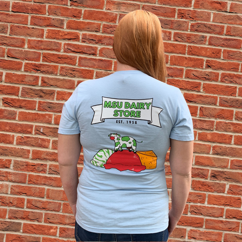 Woman with her back to camera in a blue t-shirt with a graphic that has a white ribbon with a green MSU Dairy Store in the middle, Est. 1958 underneath, a white cow with green spots standing on a red scoop of ice cream with a white and green scoop of ice cream to the left and a wedge of cheese to the right.