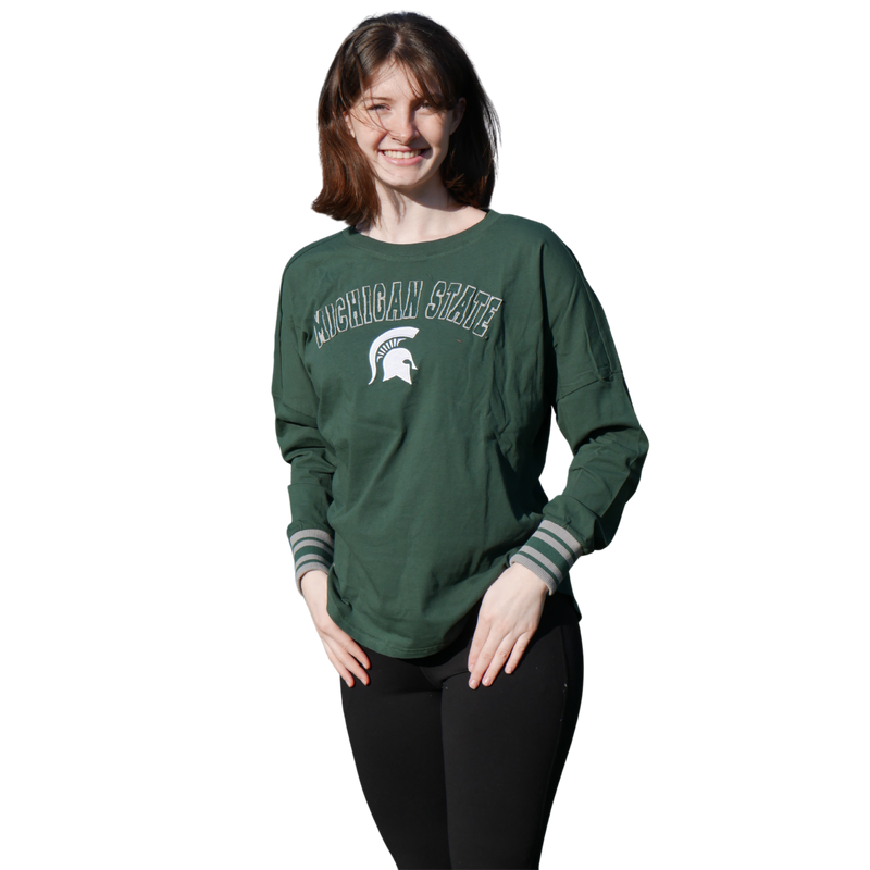 A woman wearing a green long-sleeve tee shirt. The chest has Michigan State written in all caps with a white MSU spartan helmet logo underneath. The sleeve cuffs alternate colors of green and grey.