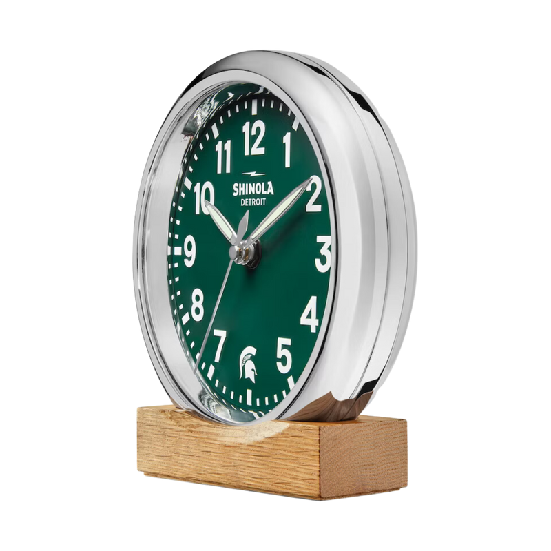 Chrome clock with green face and white lettering. Shinola Detroit displayed under the 12 and a Spartan helment in the place of the 6. White lettering for the hours and minutes, with chrome sweeping hands, on a natural oak stand.