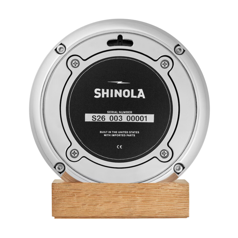 The back of a chrome clock with green face and white lettering. Shinola Detroit displayed under the 12 and a Spartan helment in the place of the 6. White lettering for the hours and minutes, with chrome sweeping hands, on a natural oak stand.