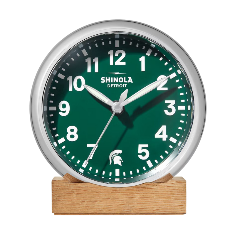 Chrome clock with green face and white lettering. Shinola Detroit displayed under the 12 and a Spartan helment in the place of the 6. White lettering for the hours and minutes, with chrome sweeping hands, on a natural oak stand.