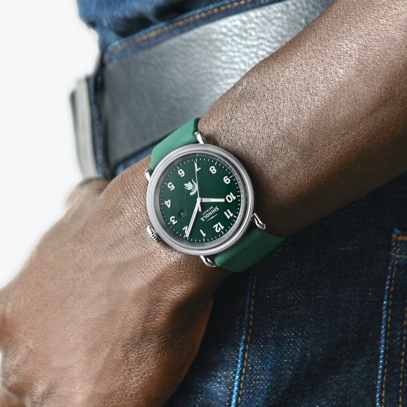A man modeling, on his wrist, a chrome watch with green face and white lettering. Shinola Detroit displayed under the 12 and a Spartan helmet above the 6. White lettering for the hours and minutes, with chrome sweeping hands, and a forest green silicone band.