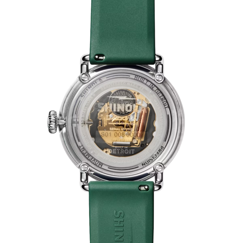 The transparent back of a chrome watch with, where the golden innerworkings can be seen through engraved glass that reads "Shinola Detroit."  The forest green silicone band is visible.