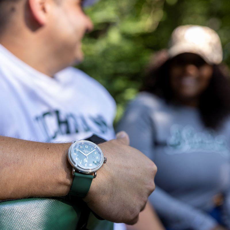 In focus is a 43 millimeter Shinola Spartan Detrola watch on a person's wrist. In the background, a man and a woman are sitting on a bench outdoors having a conversation. 