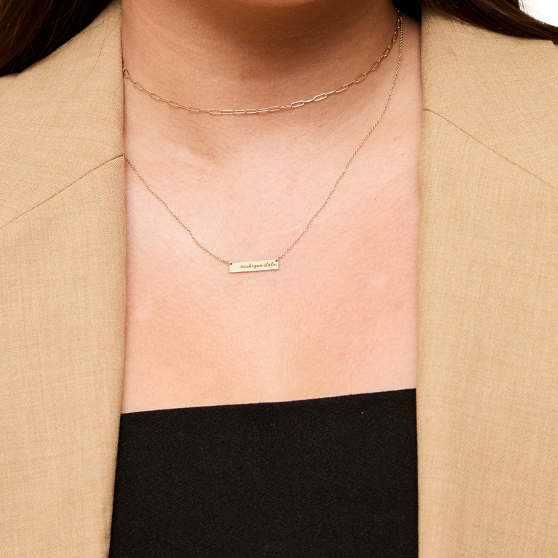 A woman wearing a gold, cable chain necklace with a silver bar at the bottom that reads Michigan State in cursive.