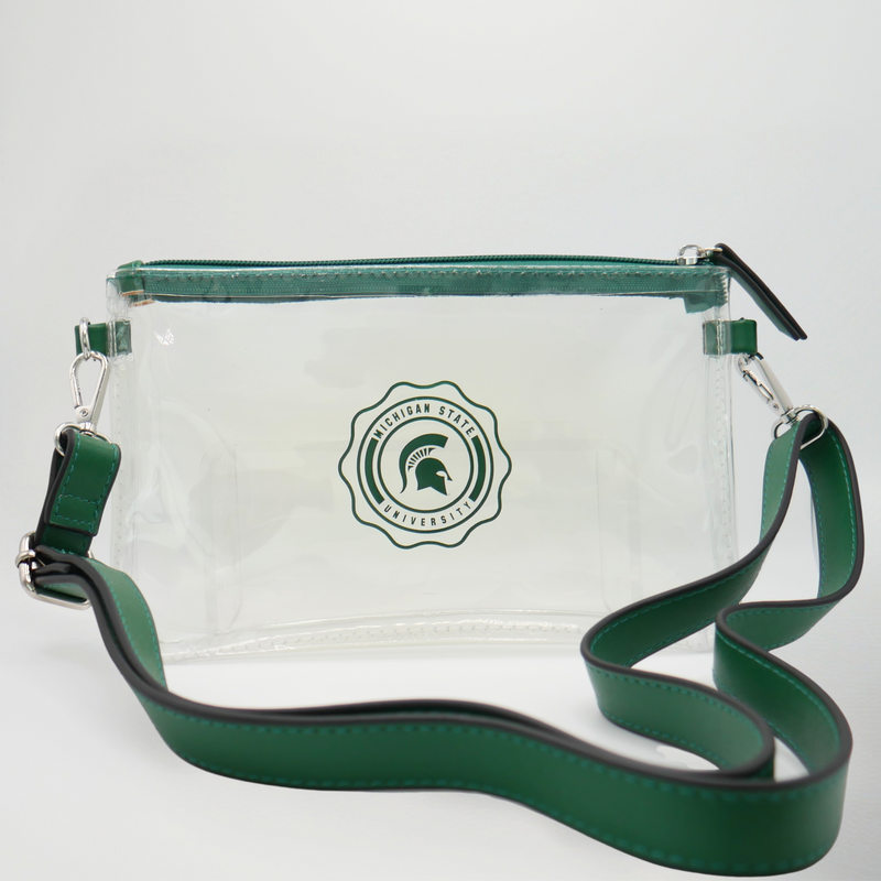 Clear rectangular pouch with green zipper. Decorative Michigan State University circle design printed on front. D-ring attachments on each side with green long crossbody strap. 